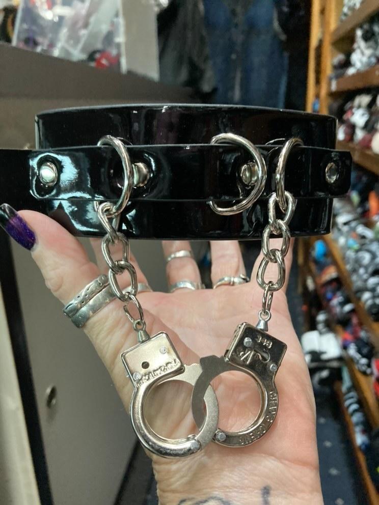 Black leather collar with silver D ring details, and two silver handcuffs hanging from two silver chains.