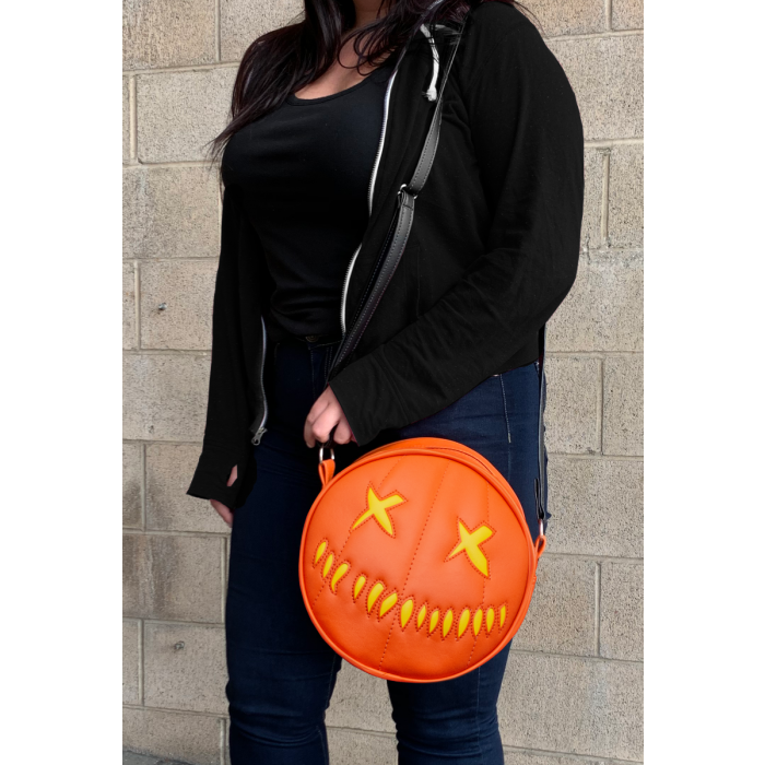 woman wearing bag on shoulder. bag is orange with yellow eyes and yellow teeth, with a black adjustable strap. based on sam's jack o lantern in the movie trick r' treat